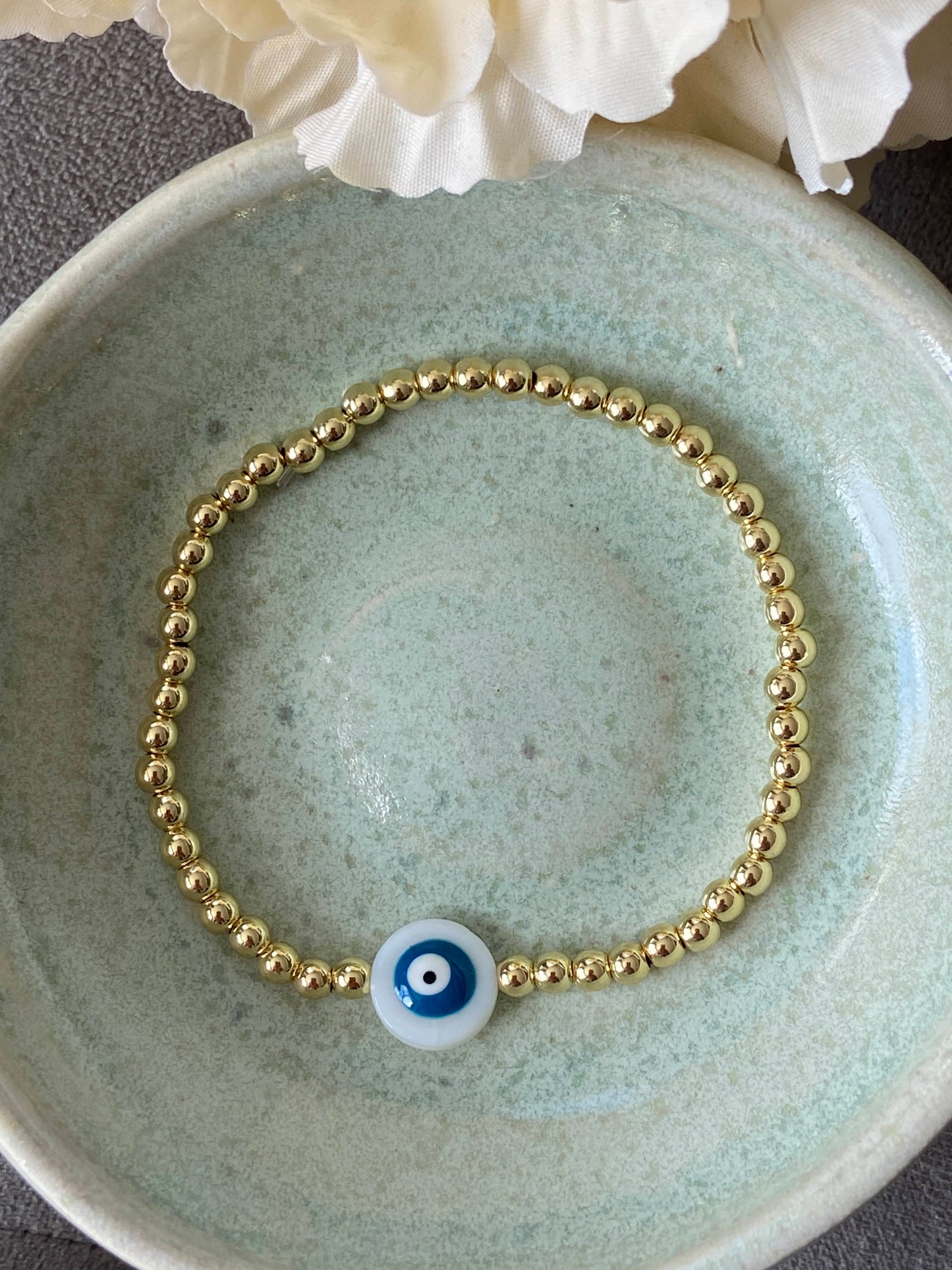 Gold bracelet with a blue evil eye charm on a ceramic dish.