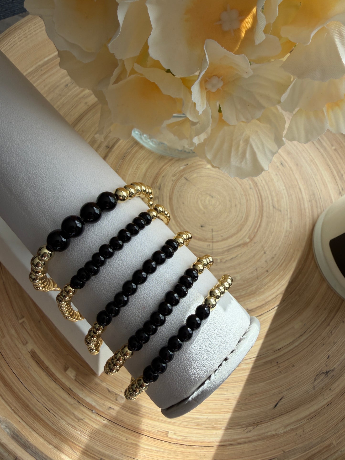 Black and gold beaded bracelets on a white cylindrical stand with flowers in the background