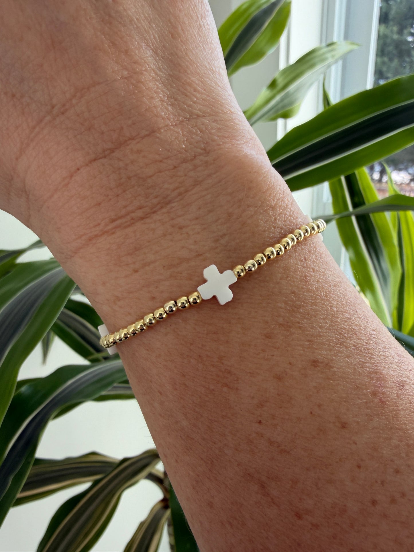 Gold bracelet with a cross charm on a wrist, with green plants in the background