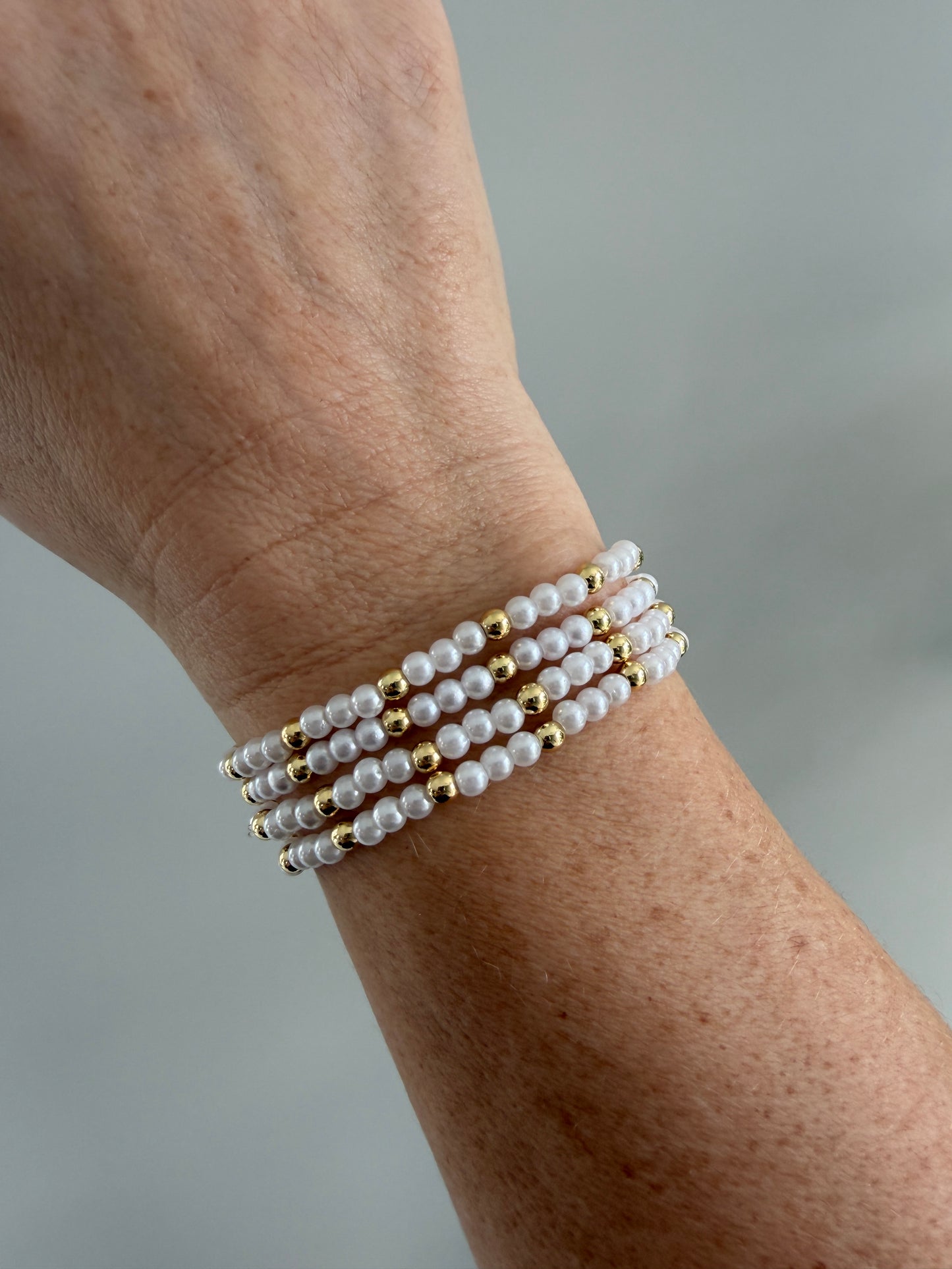 Close-up of a wrist wearing multiple pearl and gold bracelets on a neutral background