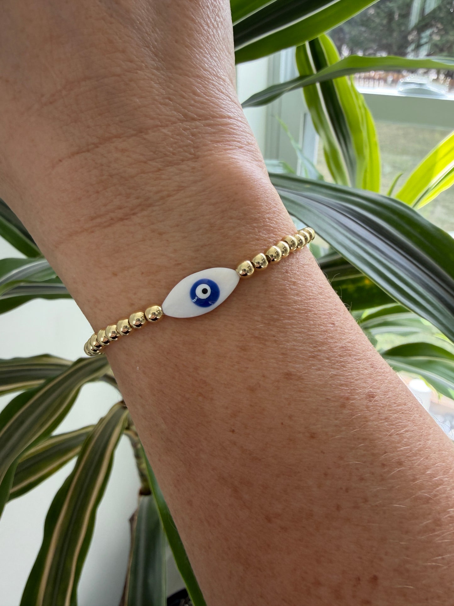 Gold bracelet with an eye design on a wrist, surrounded by green plants.