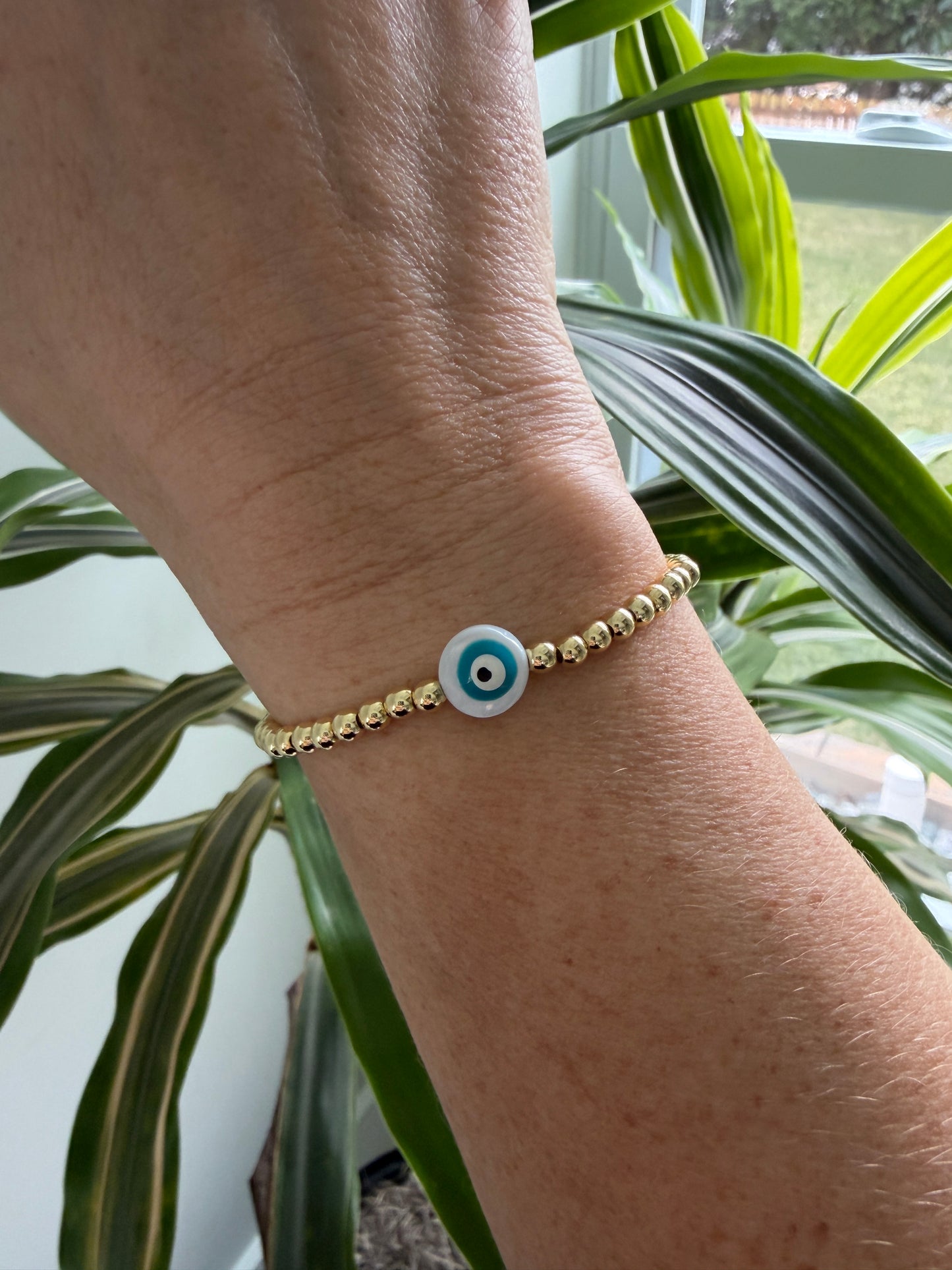 Gold bracelet with a blue evil eye charm on a wrist, with green plants in the background