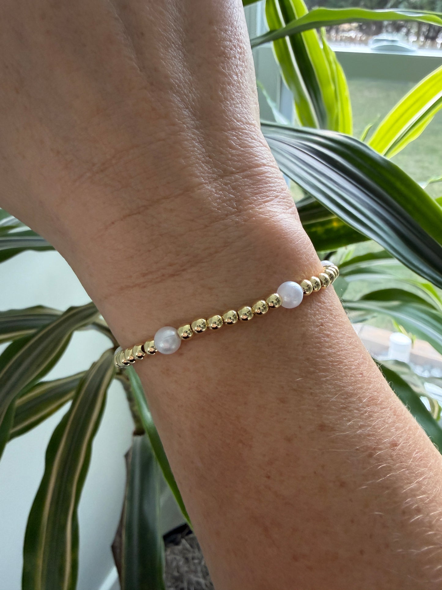 Gold beaded bracelet on a wrist with green leaves in the background