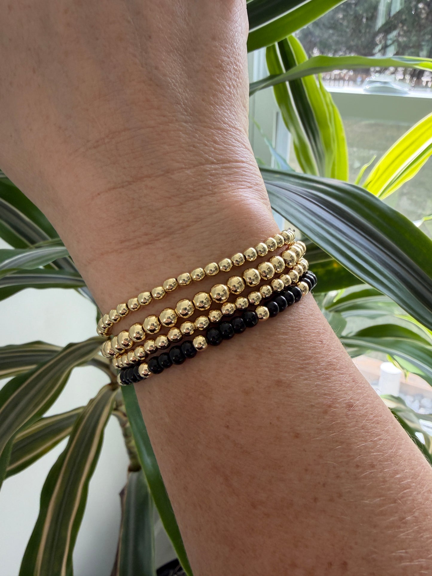 Close-up of a wrist wearing gold and black beaded bracelets with a plant in the background
