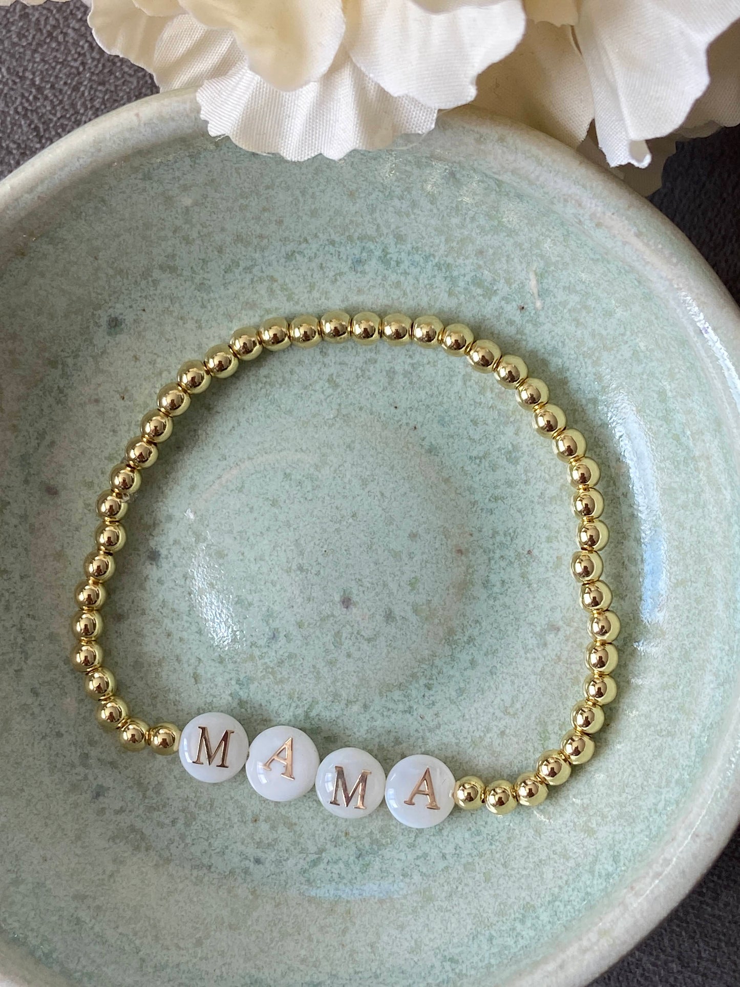 Gold bracelet with 'MAMA' beads on a ceramic dish with white flowers.