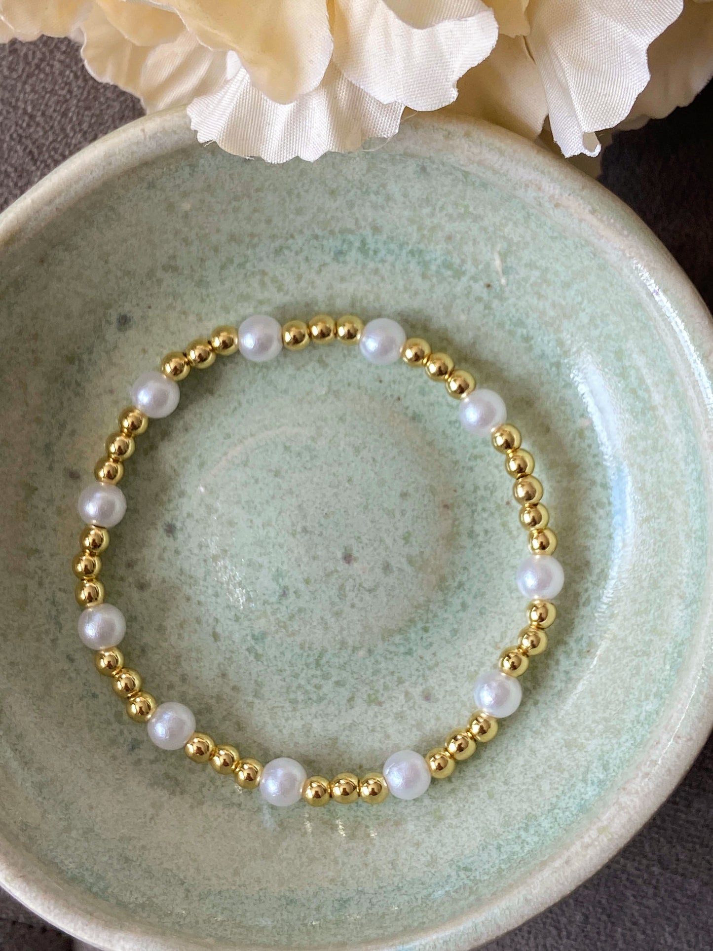 Pearl and gold beaded bracelet on a textured green plate with flowers in the background