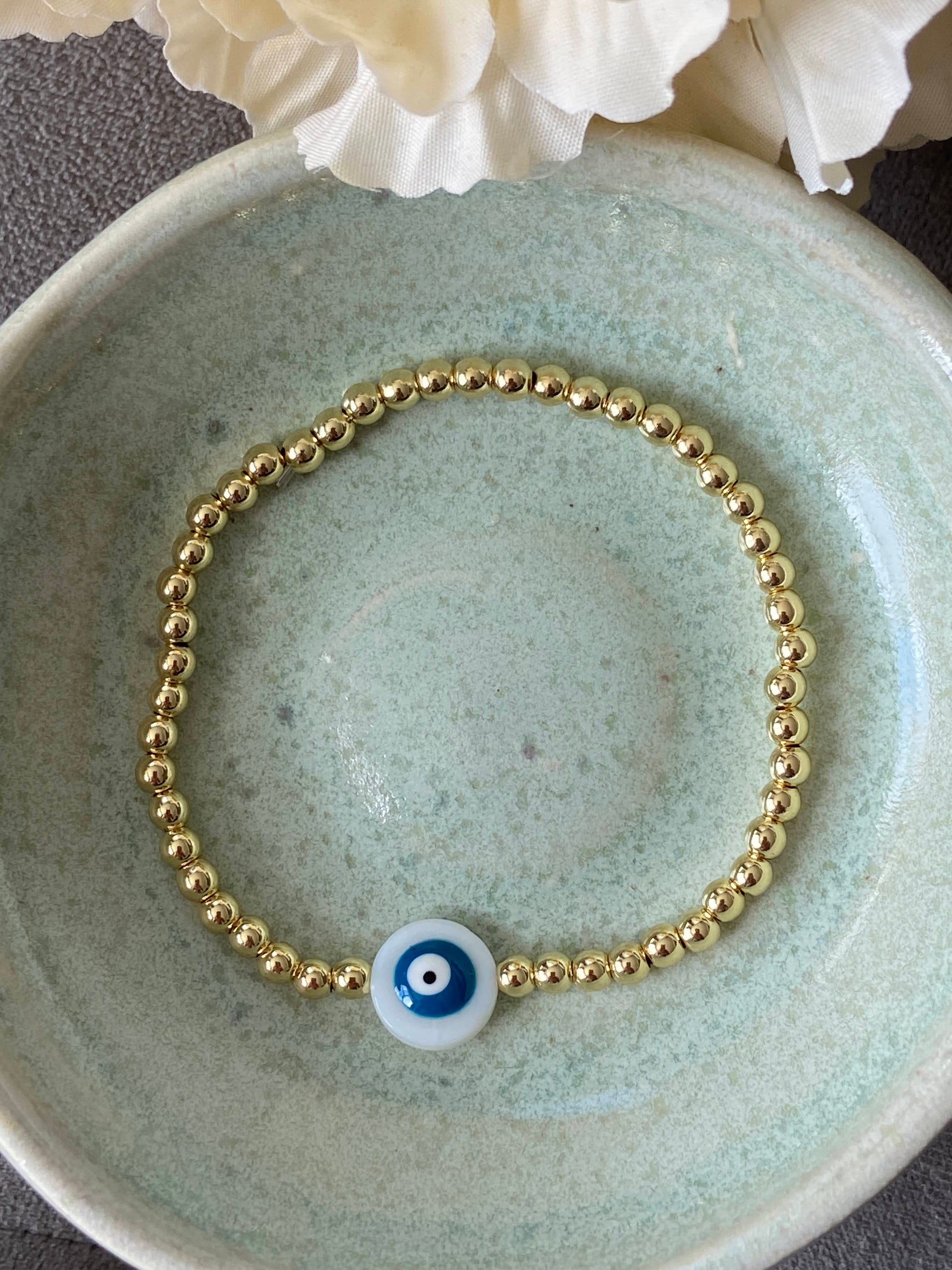 Gold bracelet with a blue evil eye charm on a ceramic dish.
