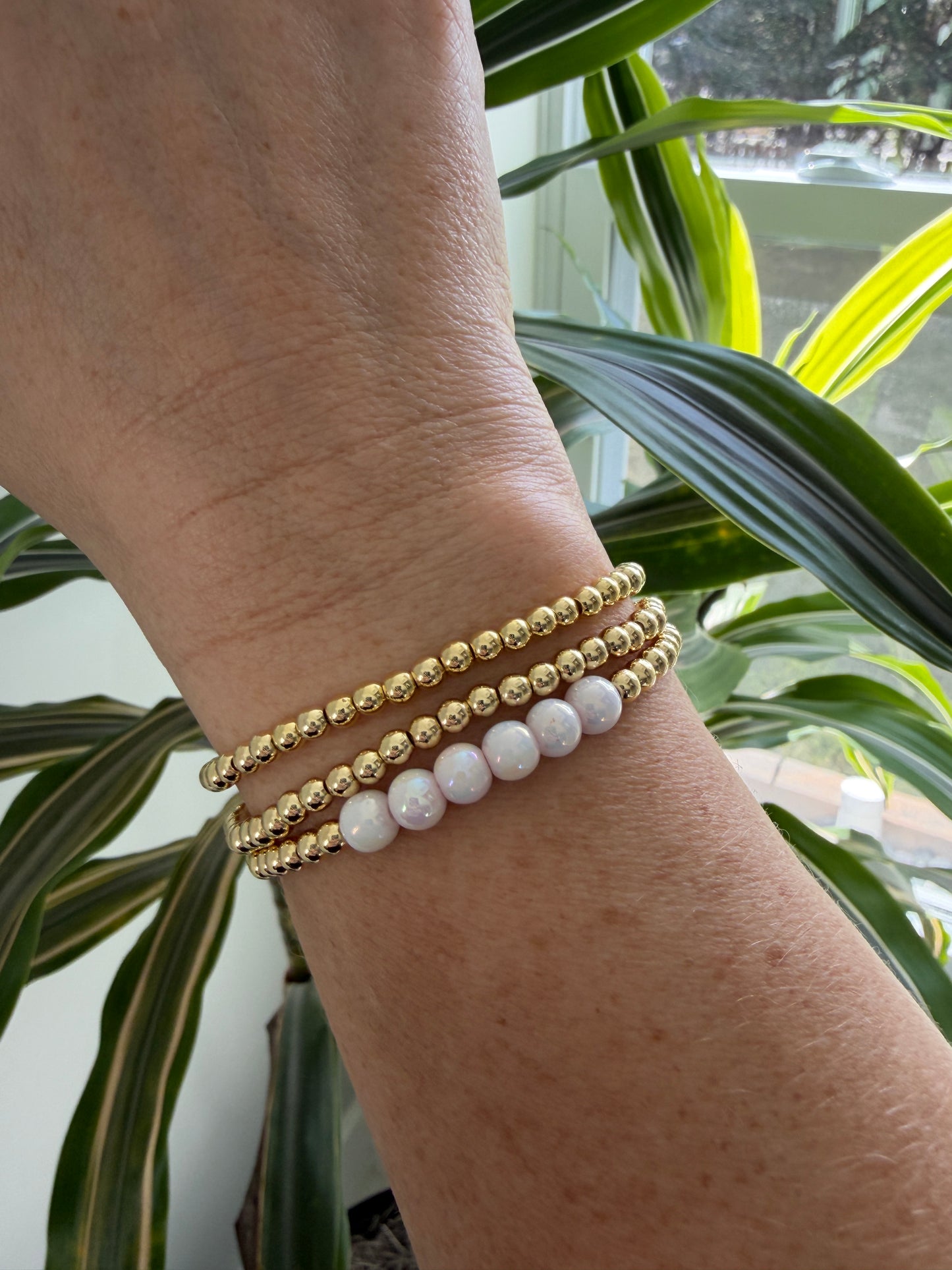 Close-up of a wrist wearing gold and white beaded bracelets with plants in the background