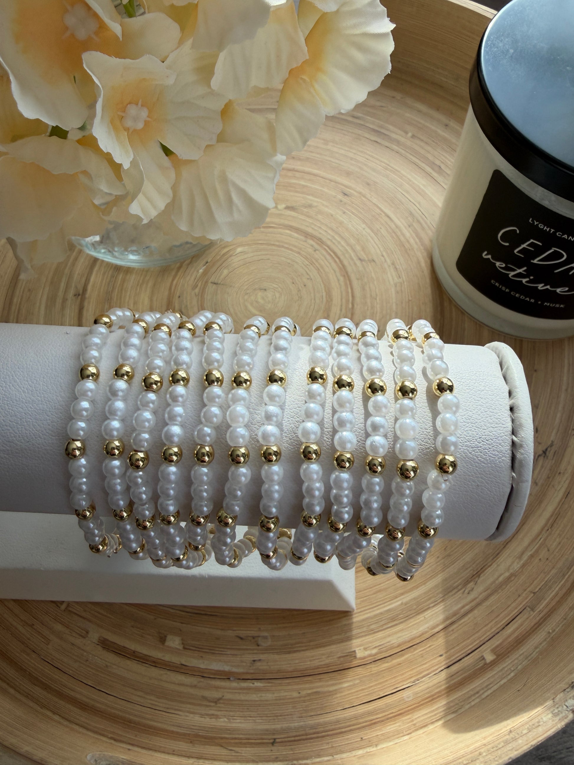 Stack of pearl bracelets with gold accents on a white stand, next to a bottle and flowers on a wooden surface.