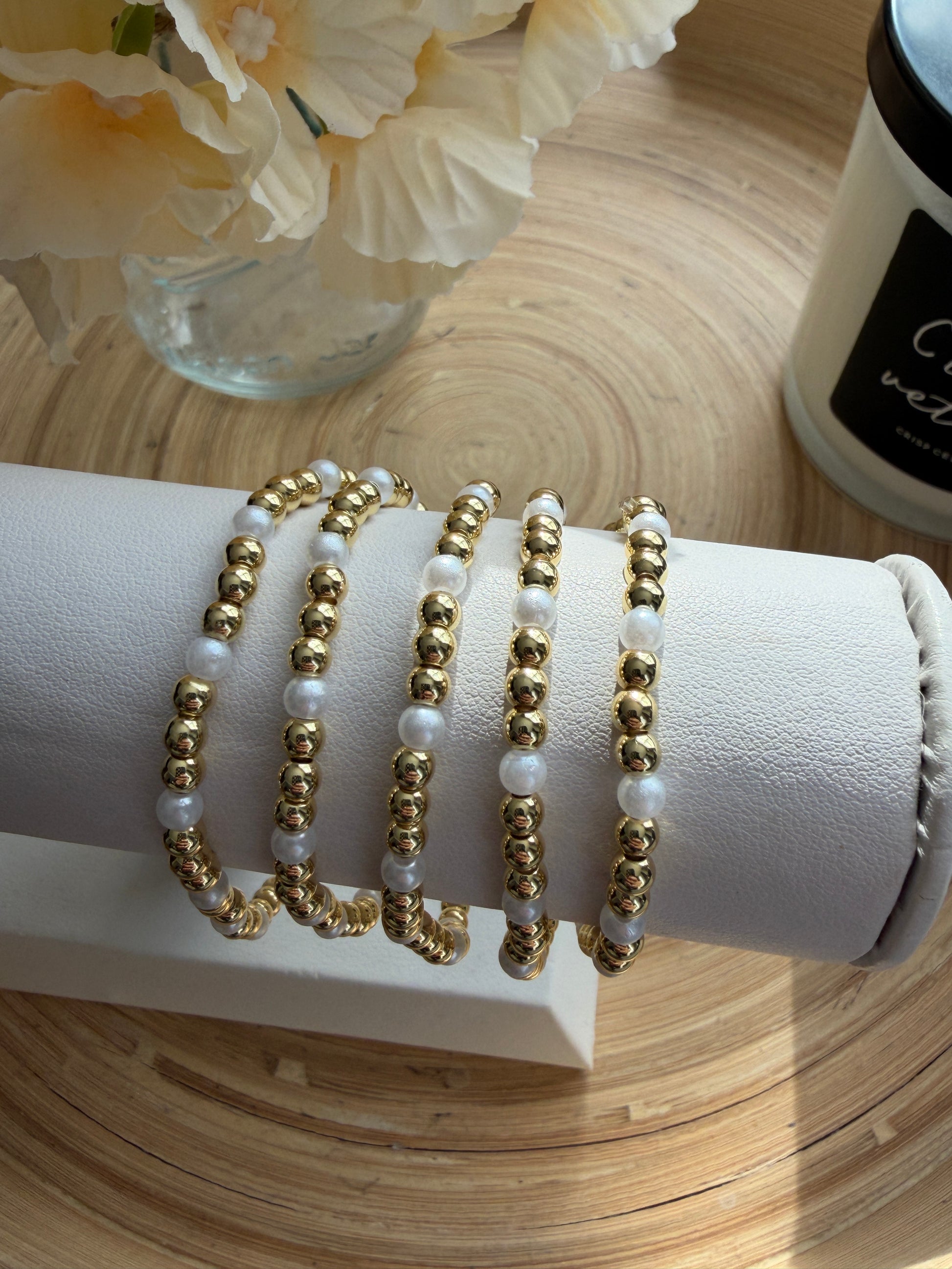 Set of gold and white beaded bracelets on a white cylindrical stand with flowers and a bottle in the background.