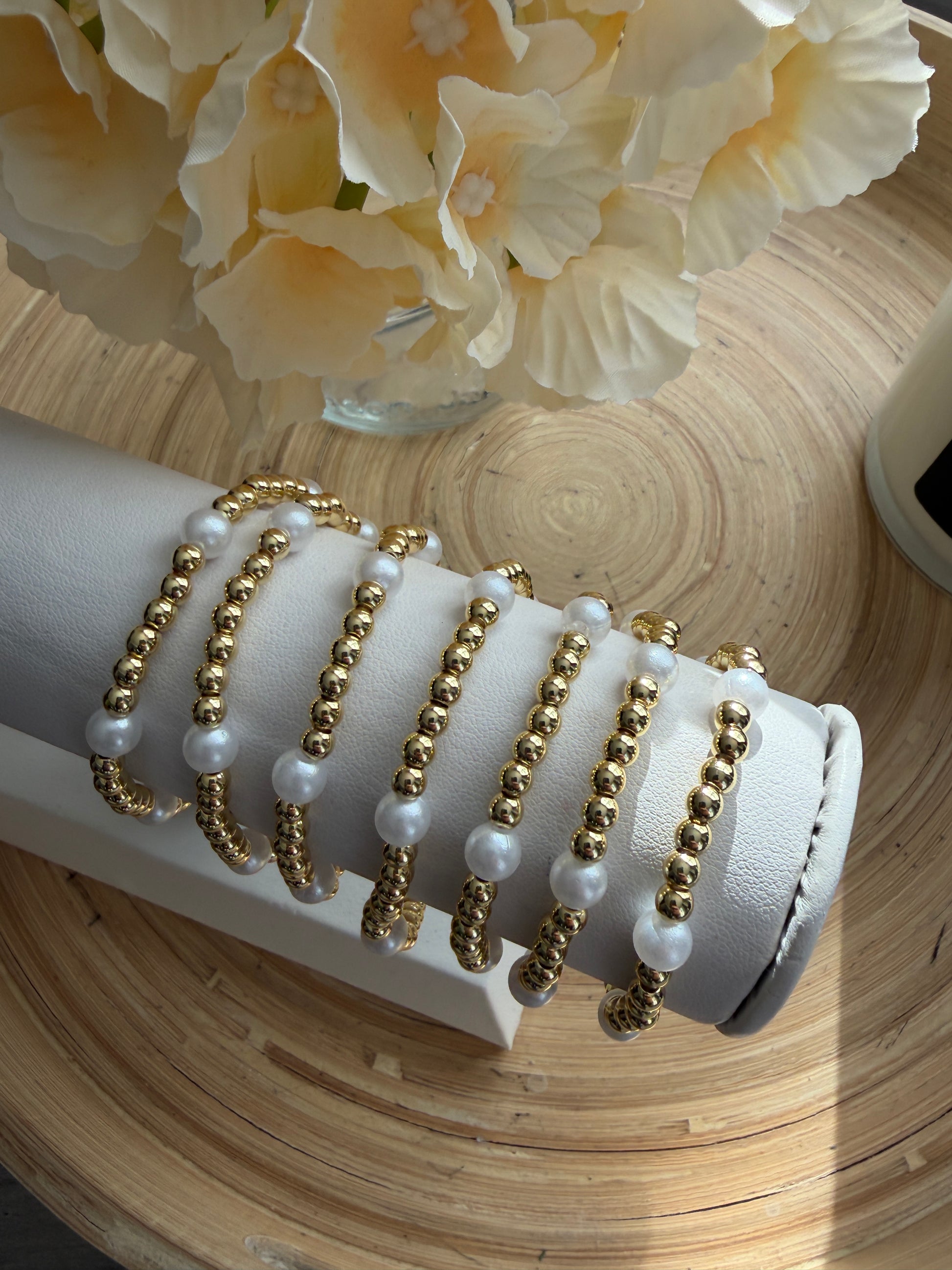 Set of gold and pearl bracelets on a white stand with flowers in the background