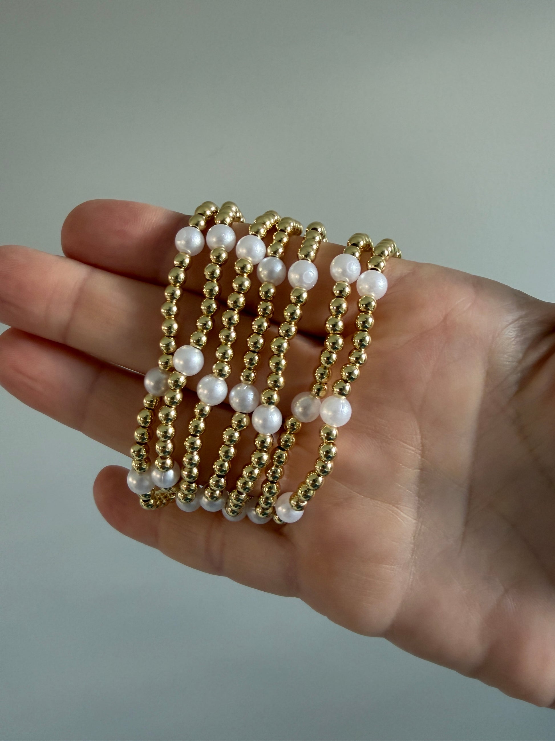 Hand wearing multiple gold and pearl beaded bracelets on a neutral background
