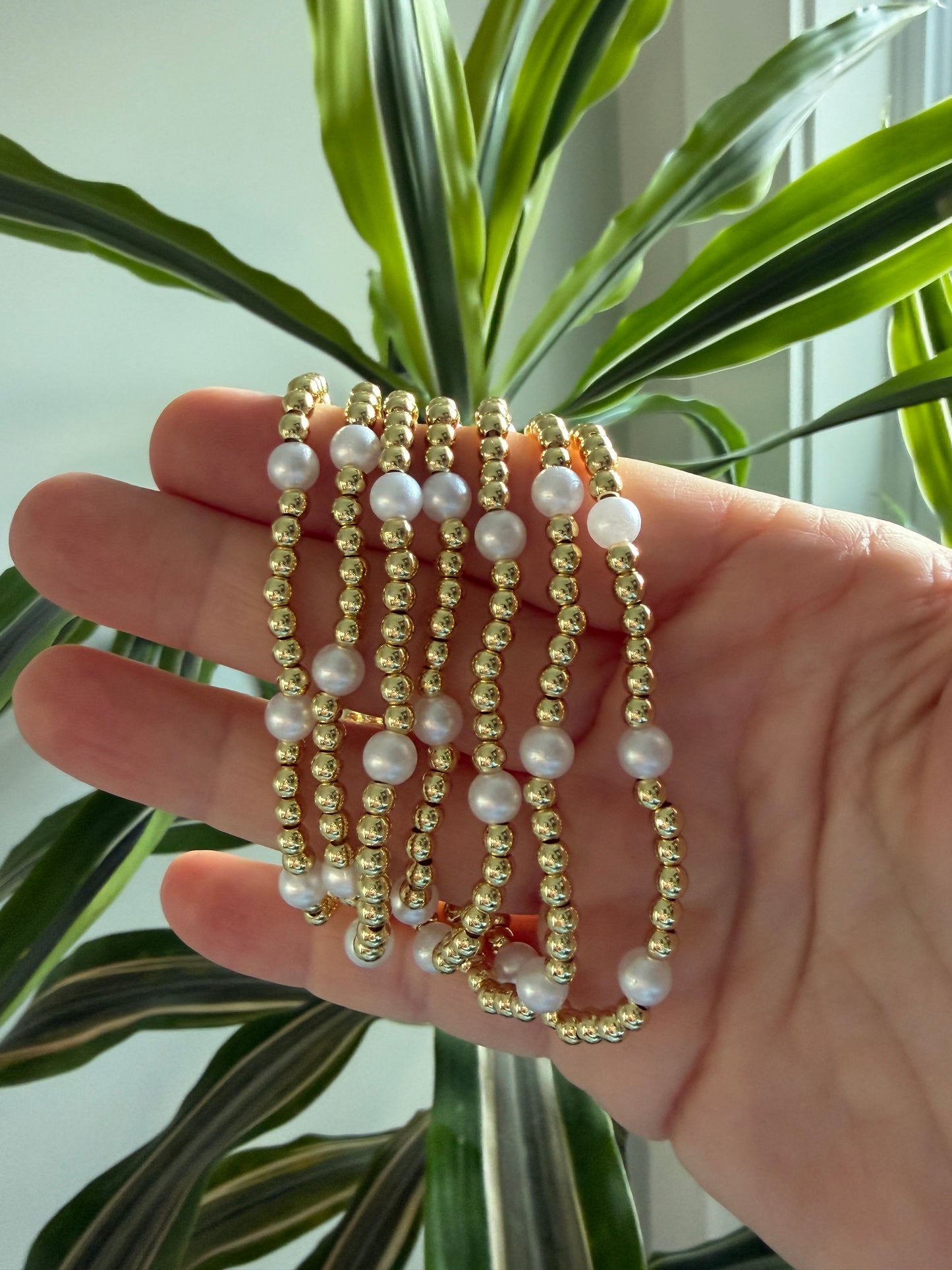 Hand wearing multiple pearl and gold beaded bracelets with a plant in the background