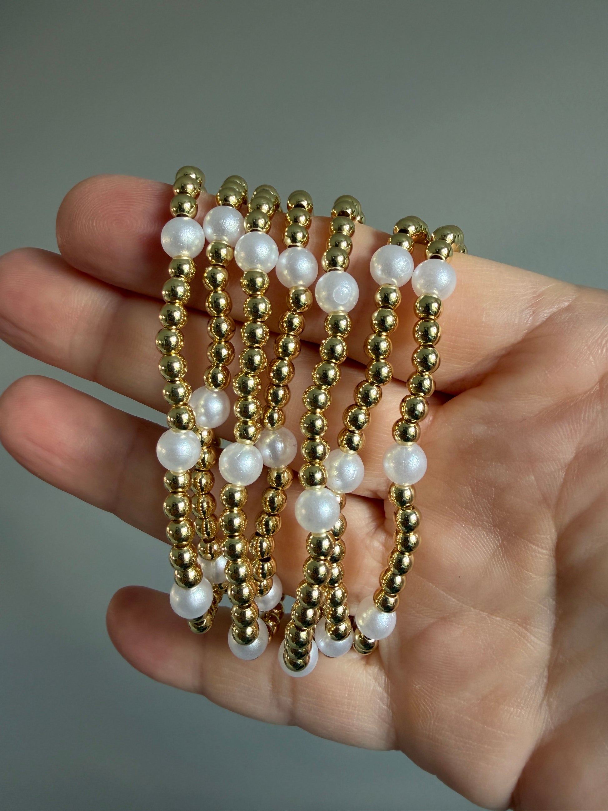 Hand holding multiple gold and pearl beaded bracelets against a neutral background