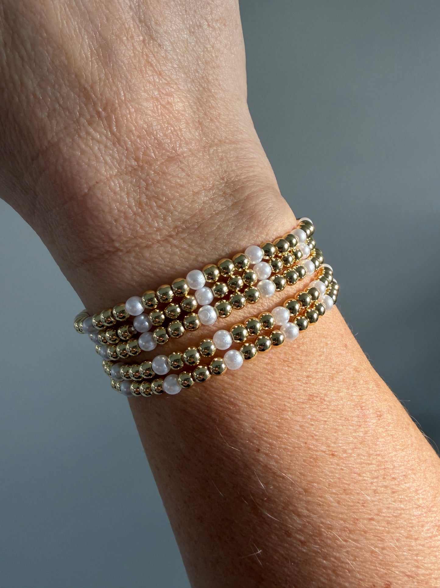 Close-up of a wrist wearing multiple gold and pearl beaded bracelets against a neutral background