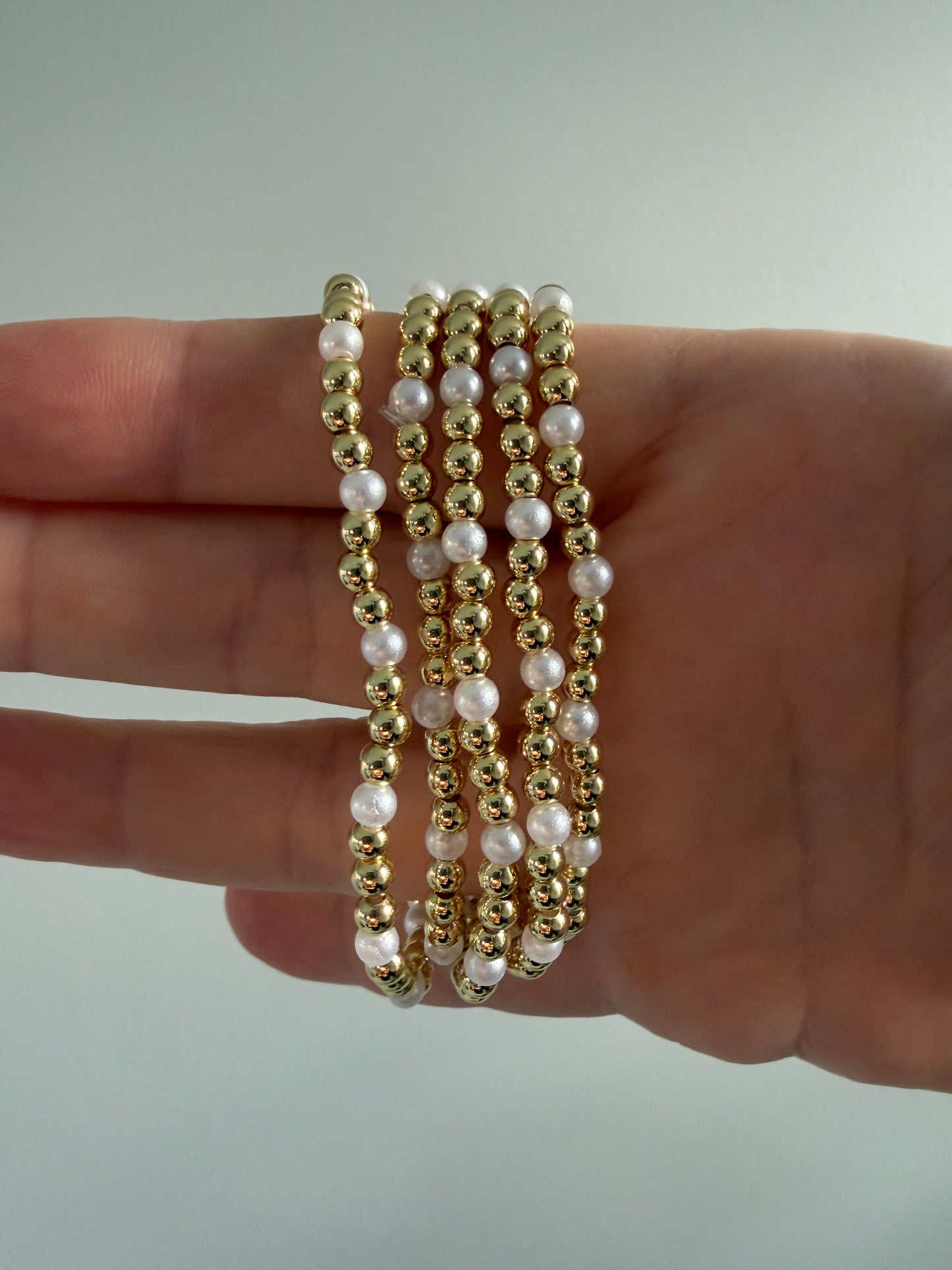 Four gold and pearl beaded bracelets on a hand against a neutral background