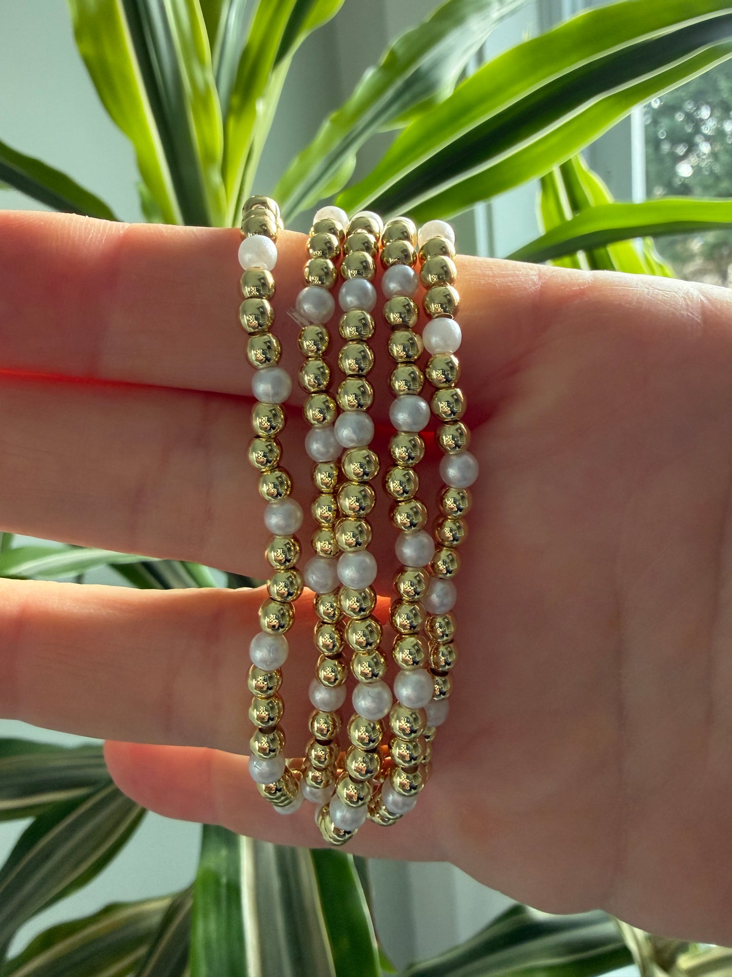 Hand wearing multiple gold and pearl beaded bracelets with a plant in the background