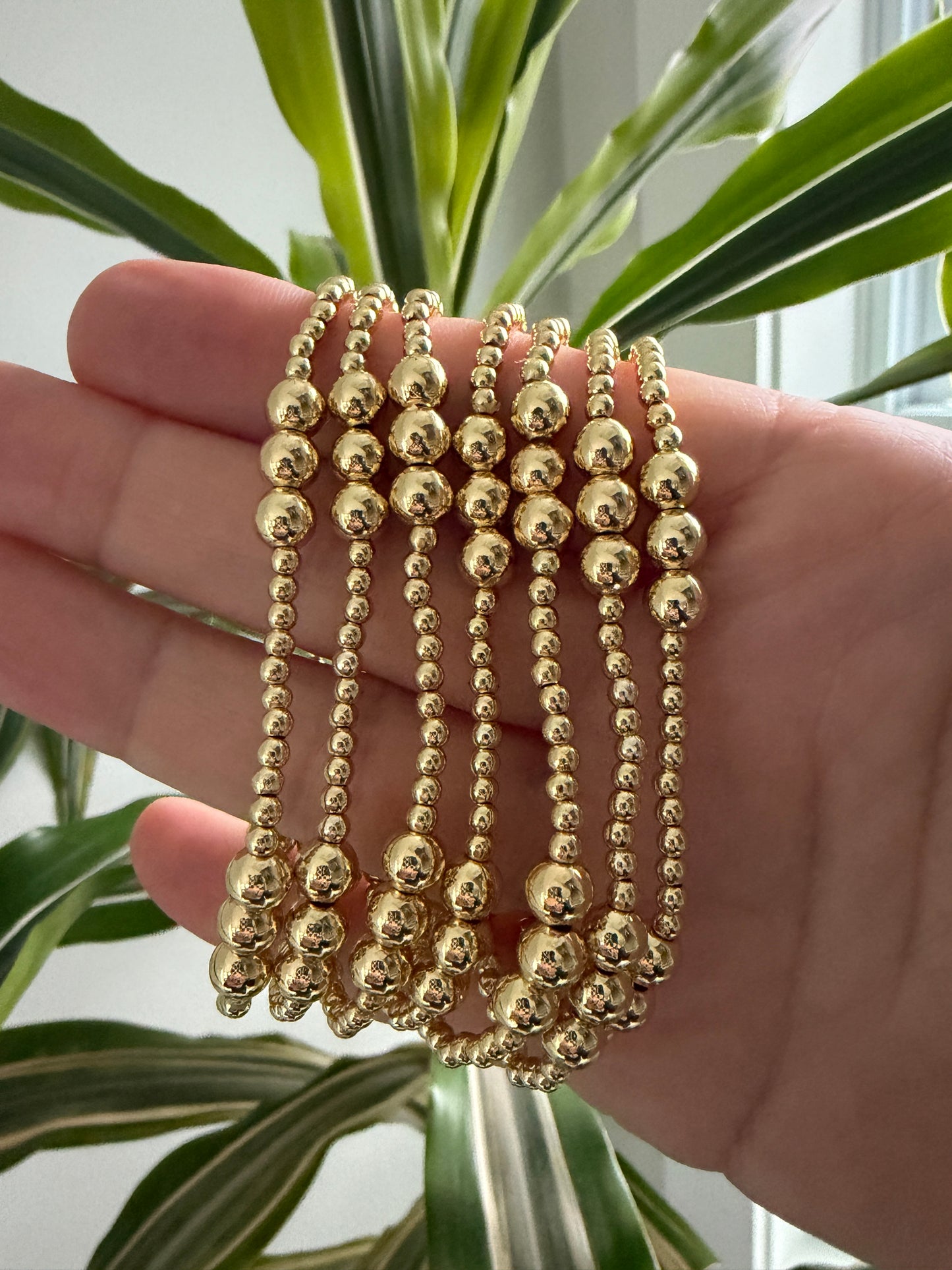 Hand wearing multiple gold beaded bracelets with a plant in the background