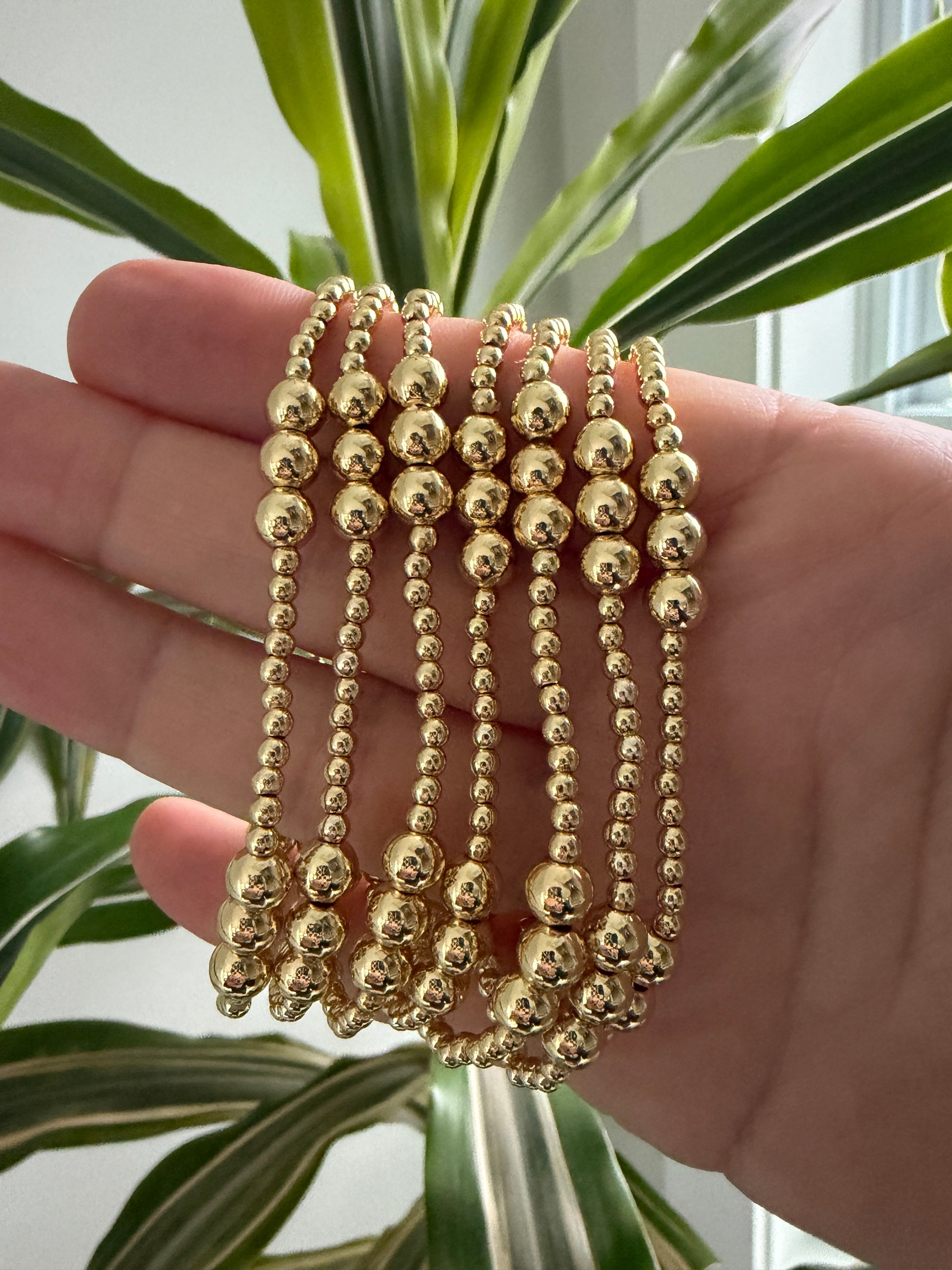 Hand wearing multiple gold beaded bracelets with a plant in the background