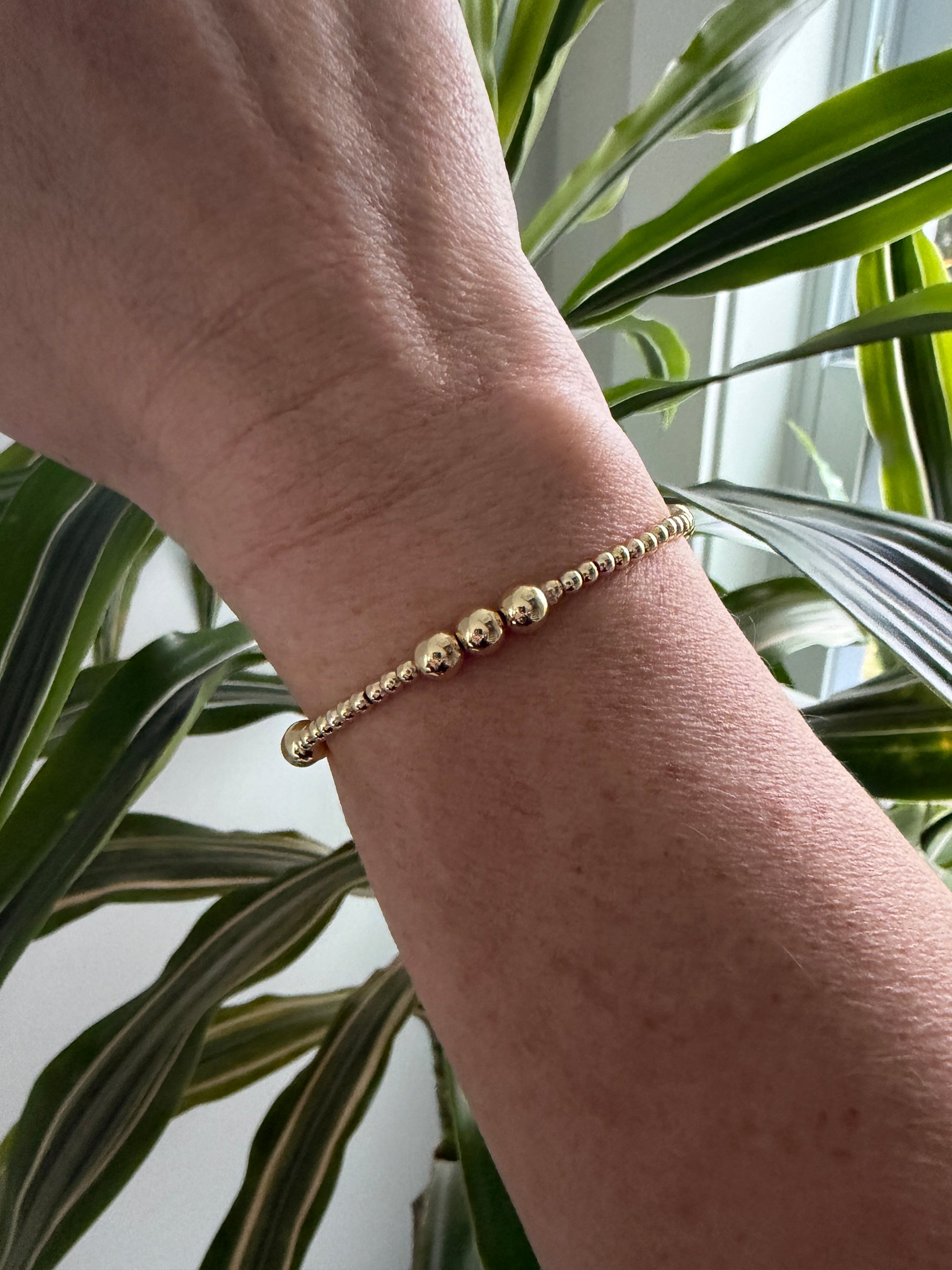 Gold beaded bracelet on a wrist with a plant in the background