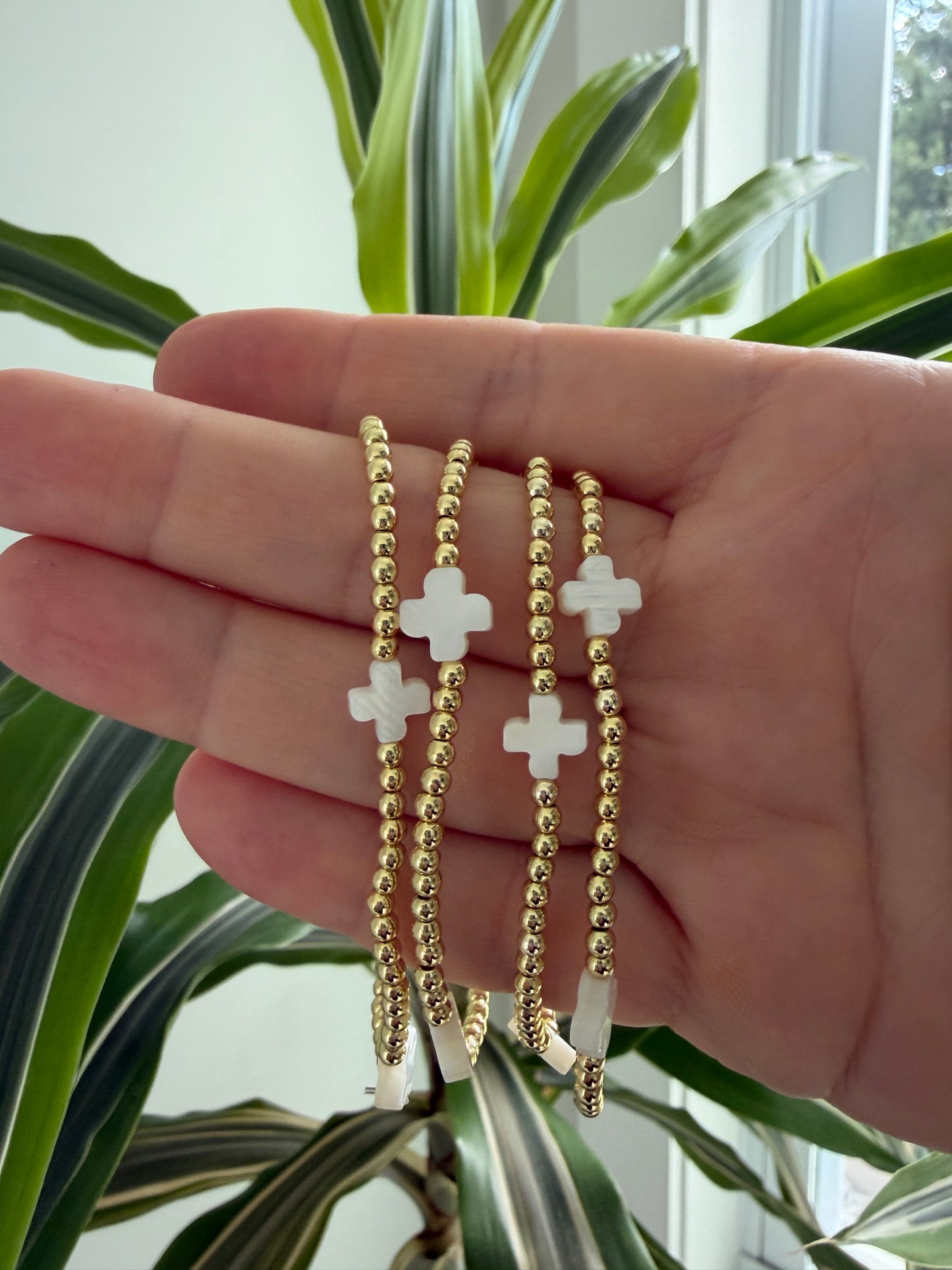 Hand wearing three gold and white cross bracelets with a plant in the background
