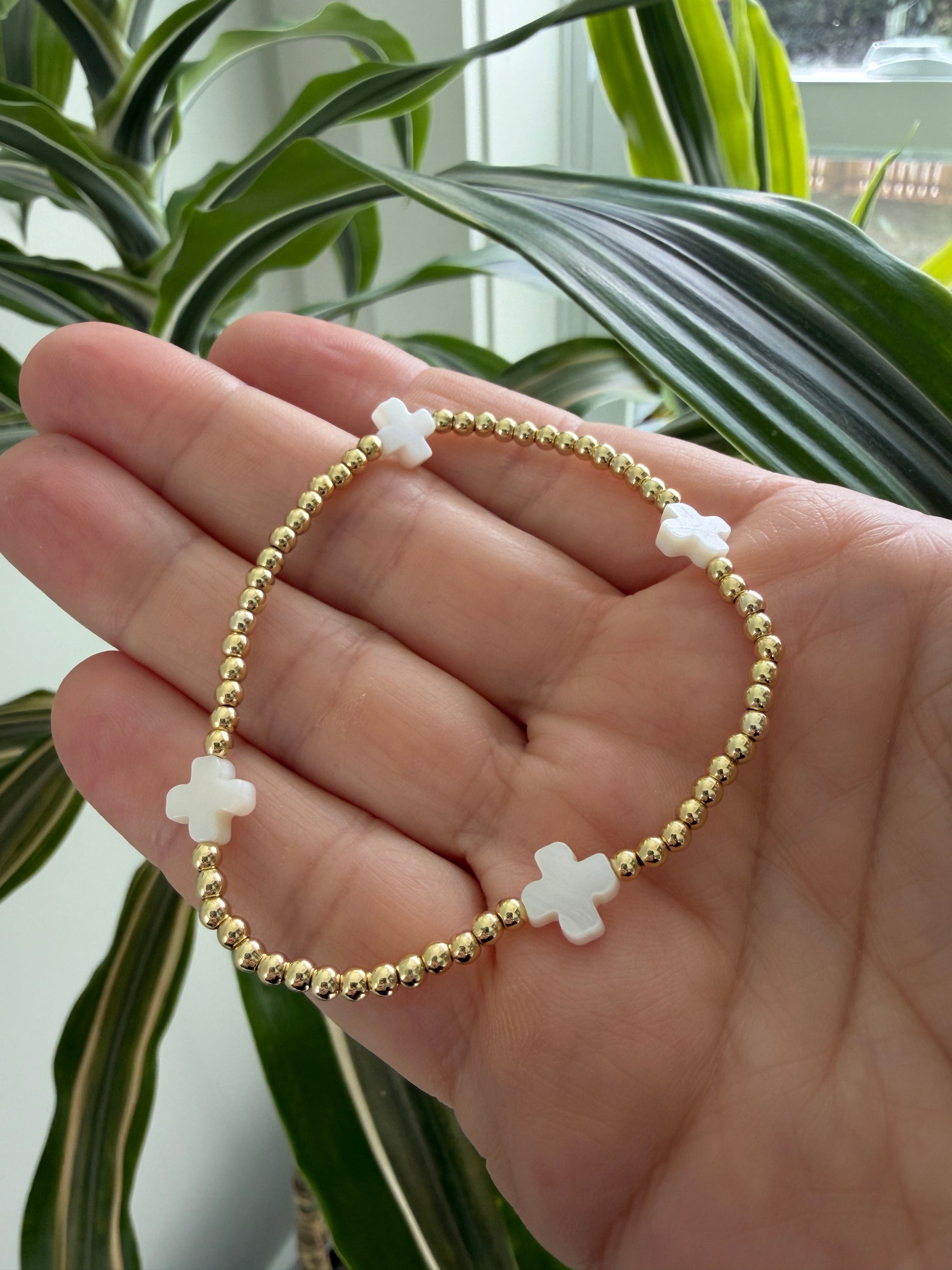 Gold bracelet with white cross charms on a hand, with green leaves in the background