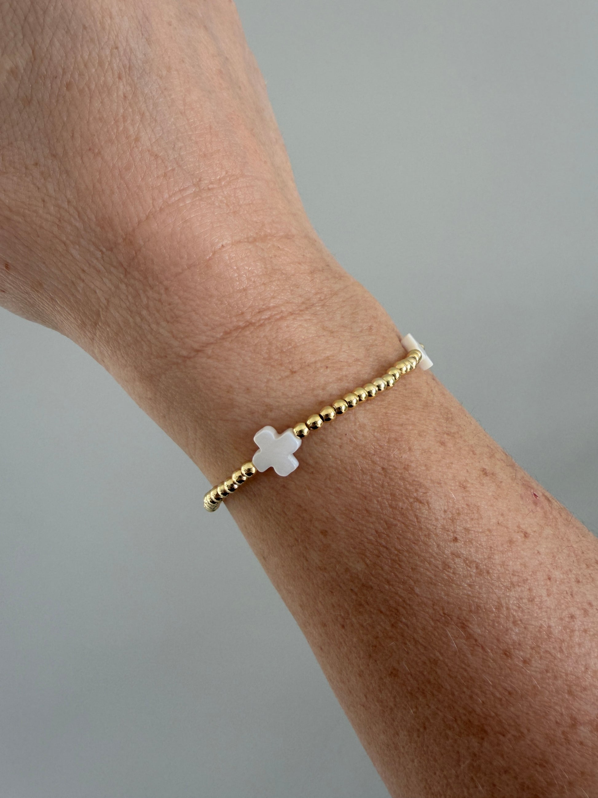 Gold bracelet with a white cross charm on a wrist against a gray background