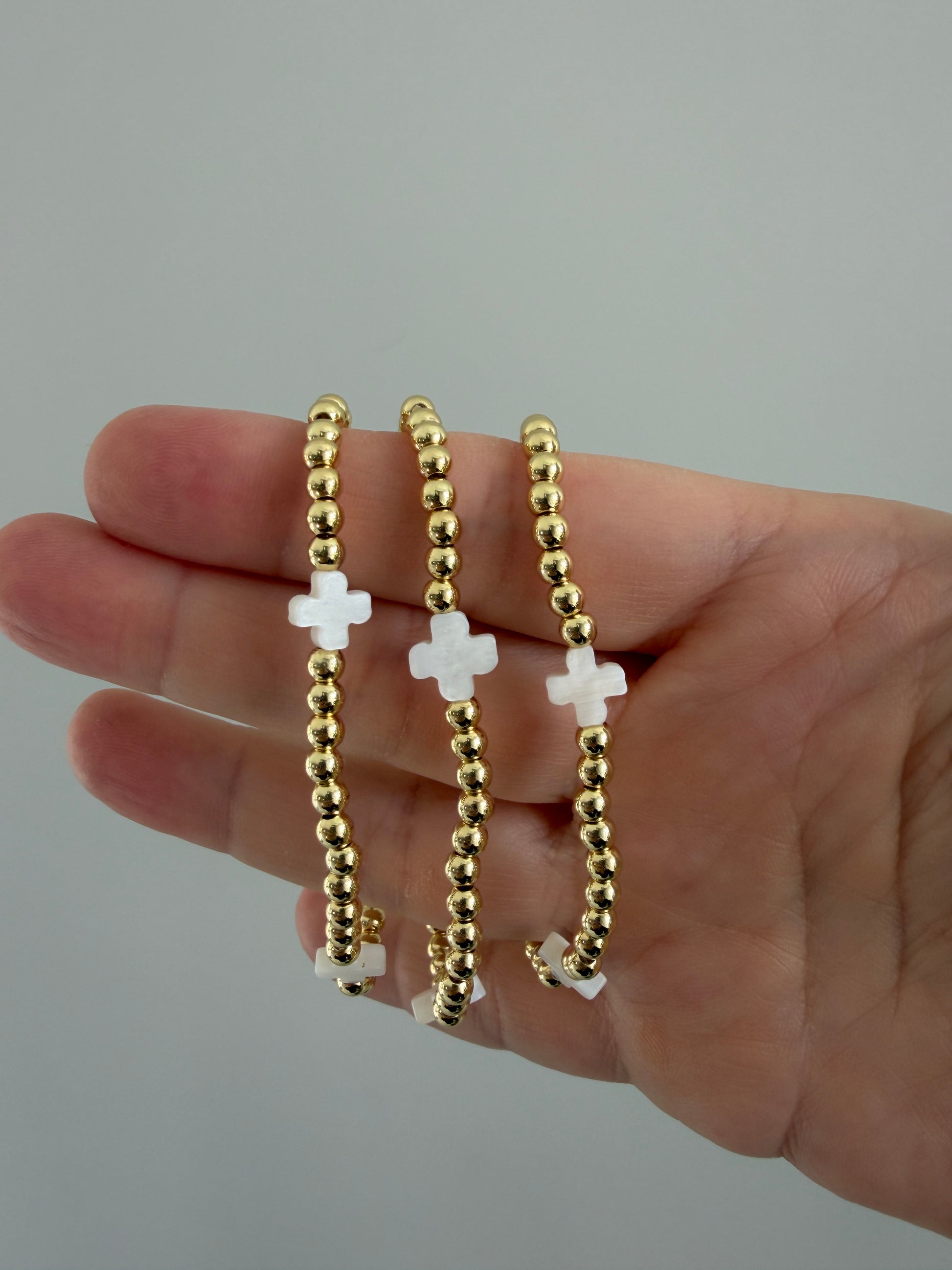 Gold beaded bracelets with white crosses on a hand against a gray background