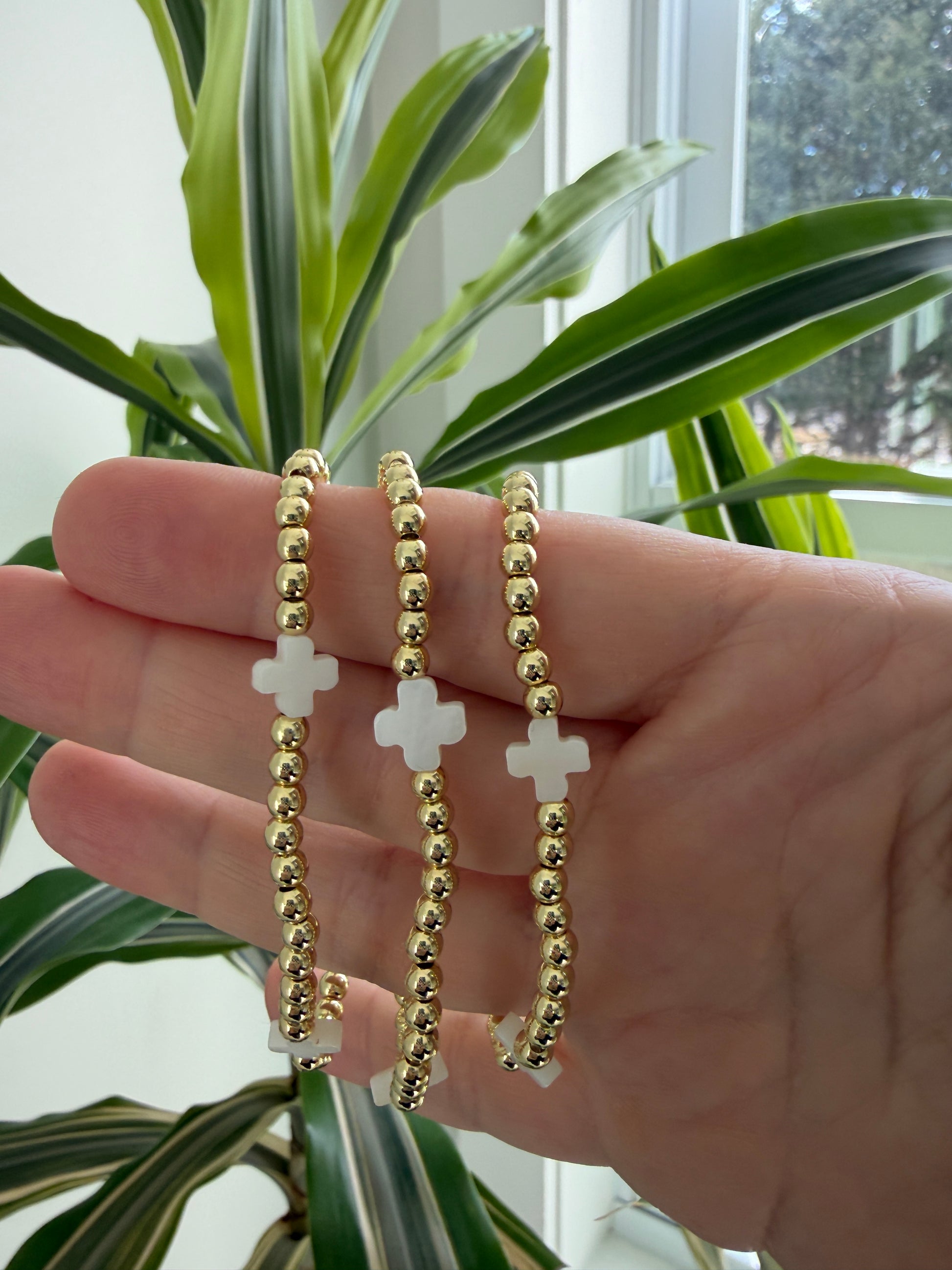 Hand wearing three gold beaded bracelets with white crosses, against a plant background