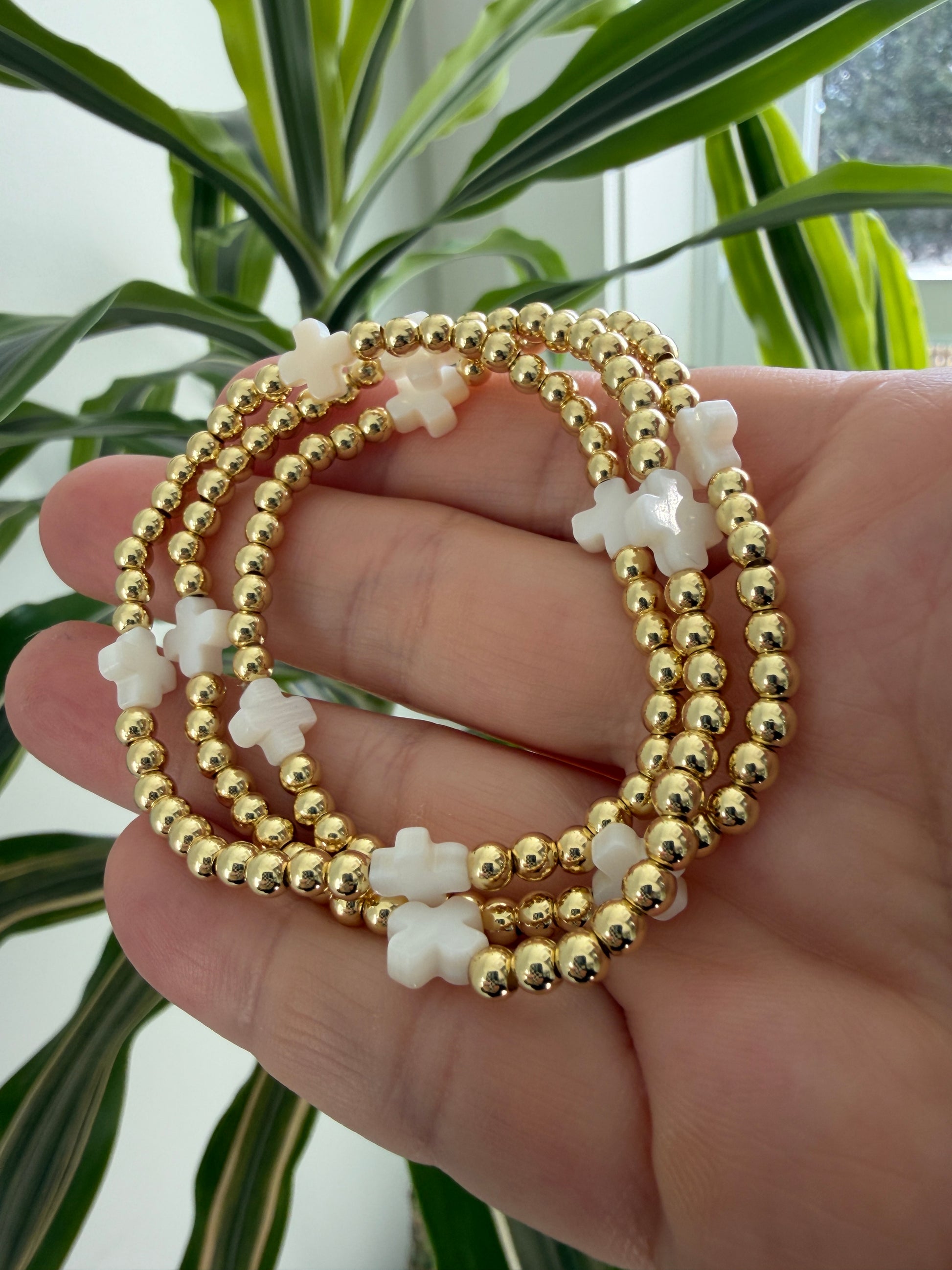 Gold beaded bracelets with white cross beads on a hand, with green plants in the background