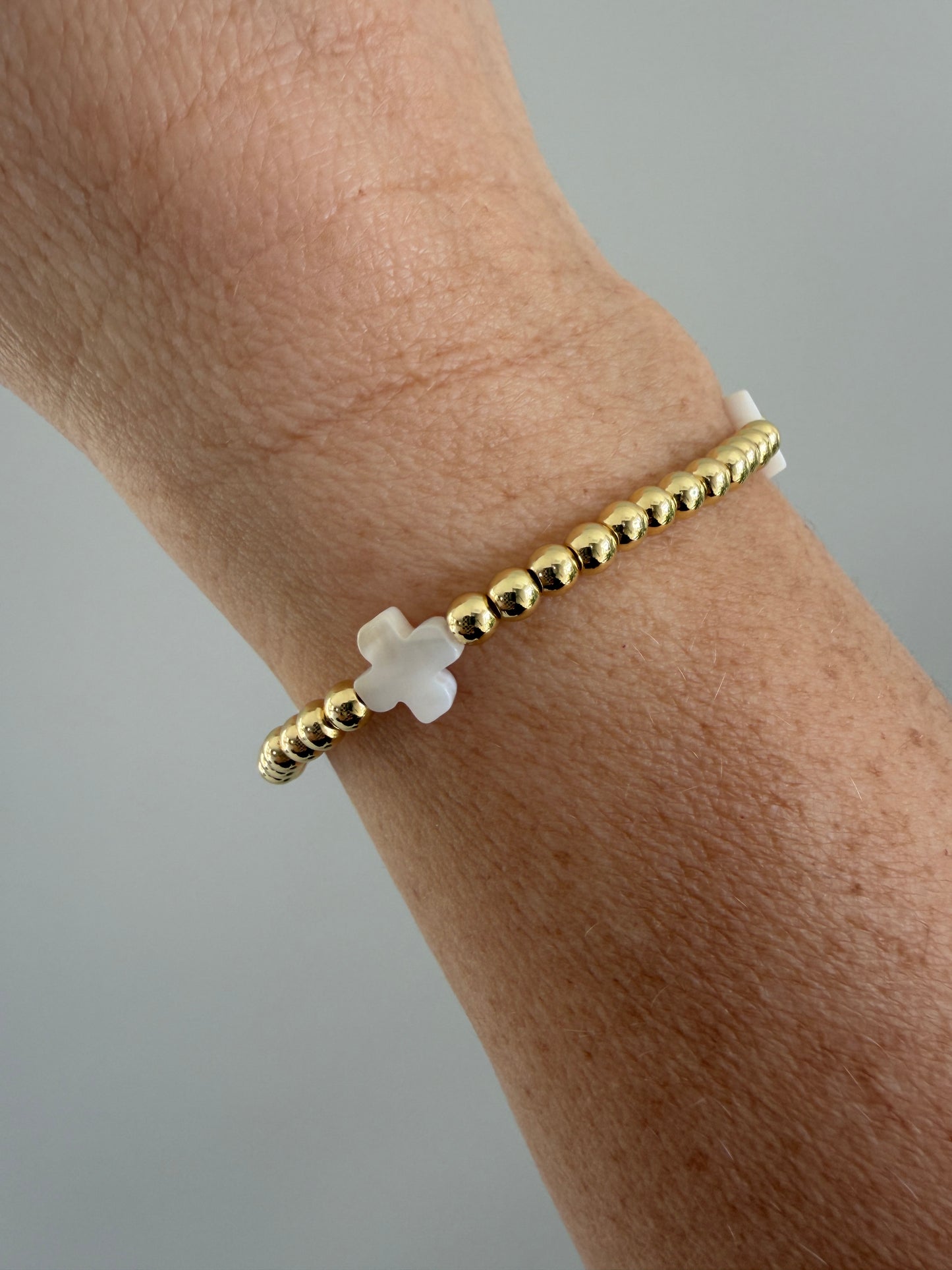 Gold beaded bracelet with a white cross charm on a wrist against a gray background