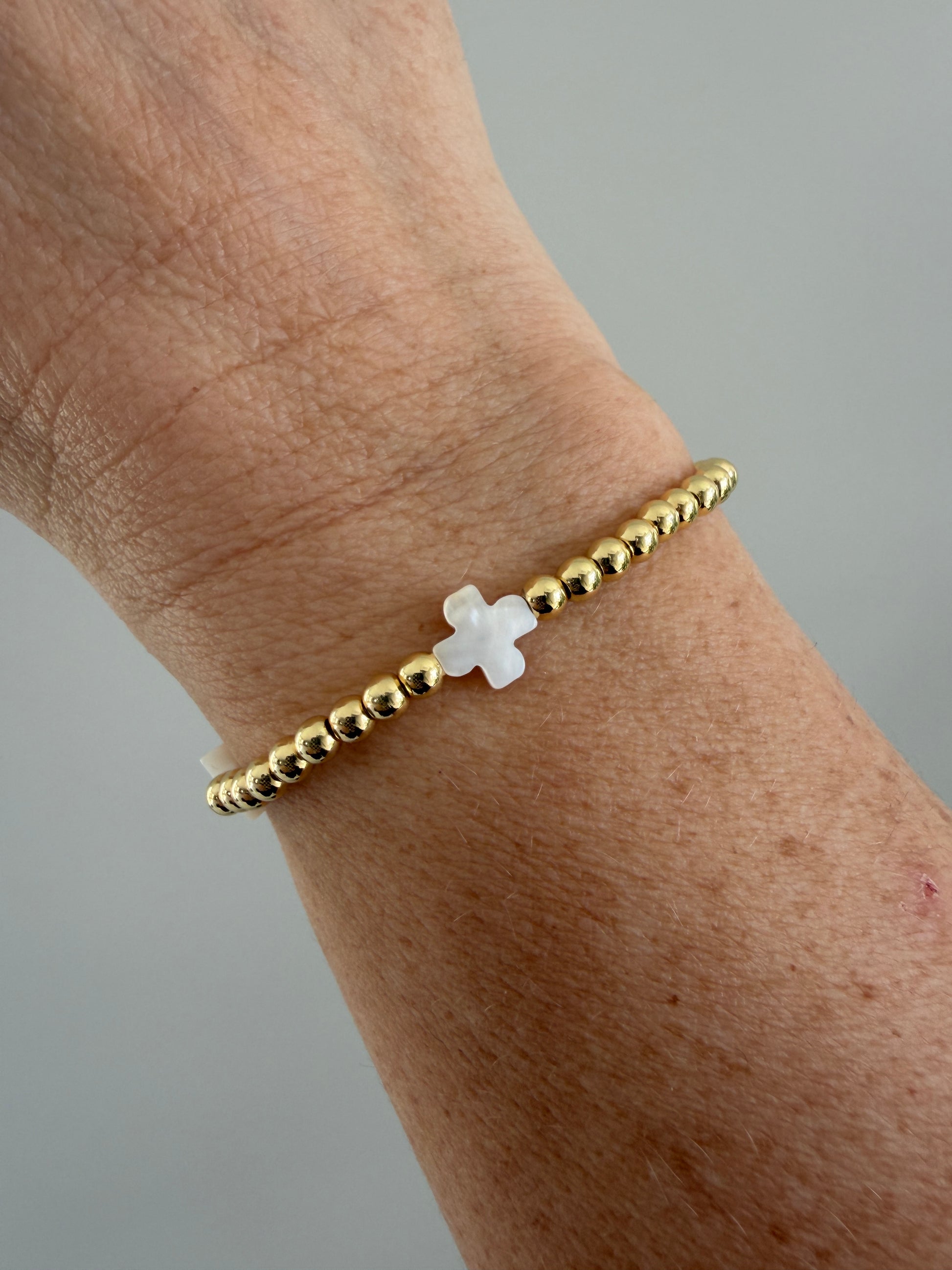 Gold beaded bracelet with a white cross on a wrist against a gray background