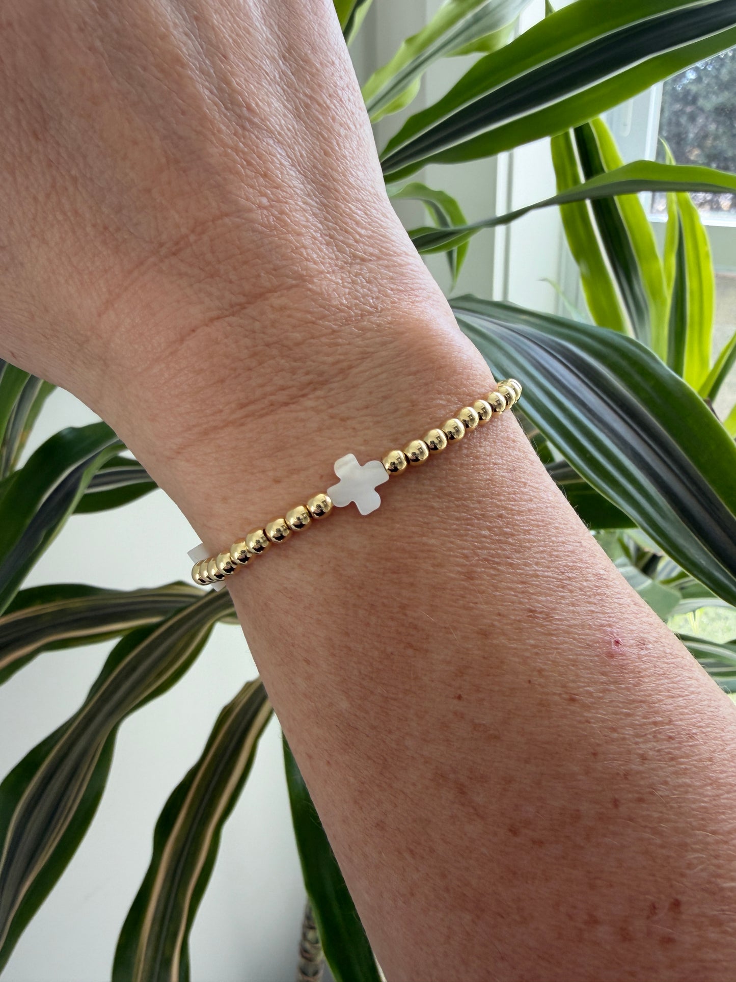 Gold bracelet with a white cross on a wrist in front of green plants