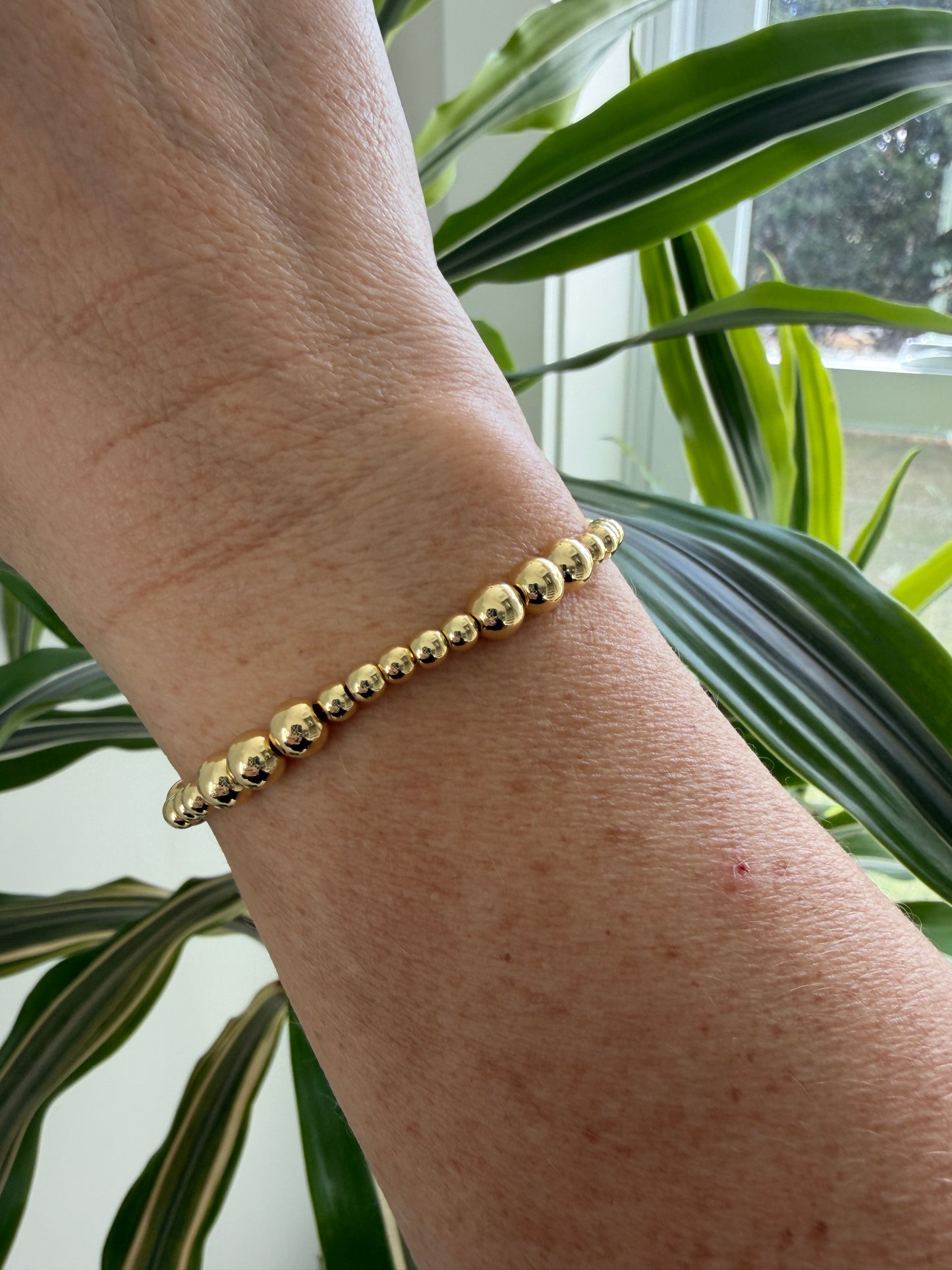 Gold beaded bracelet on a wrist with green plants in the background