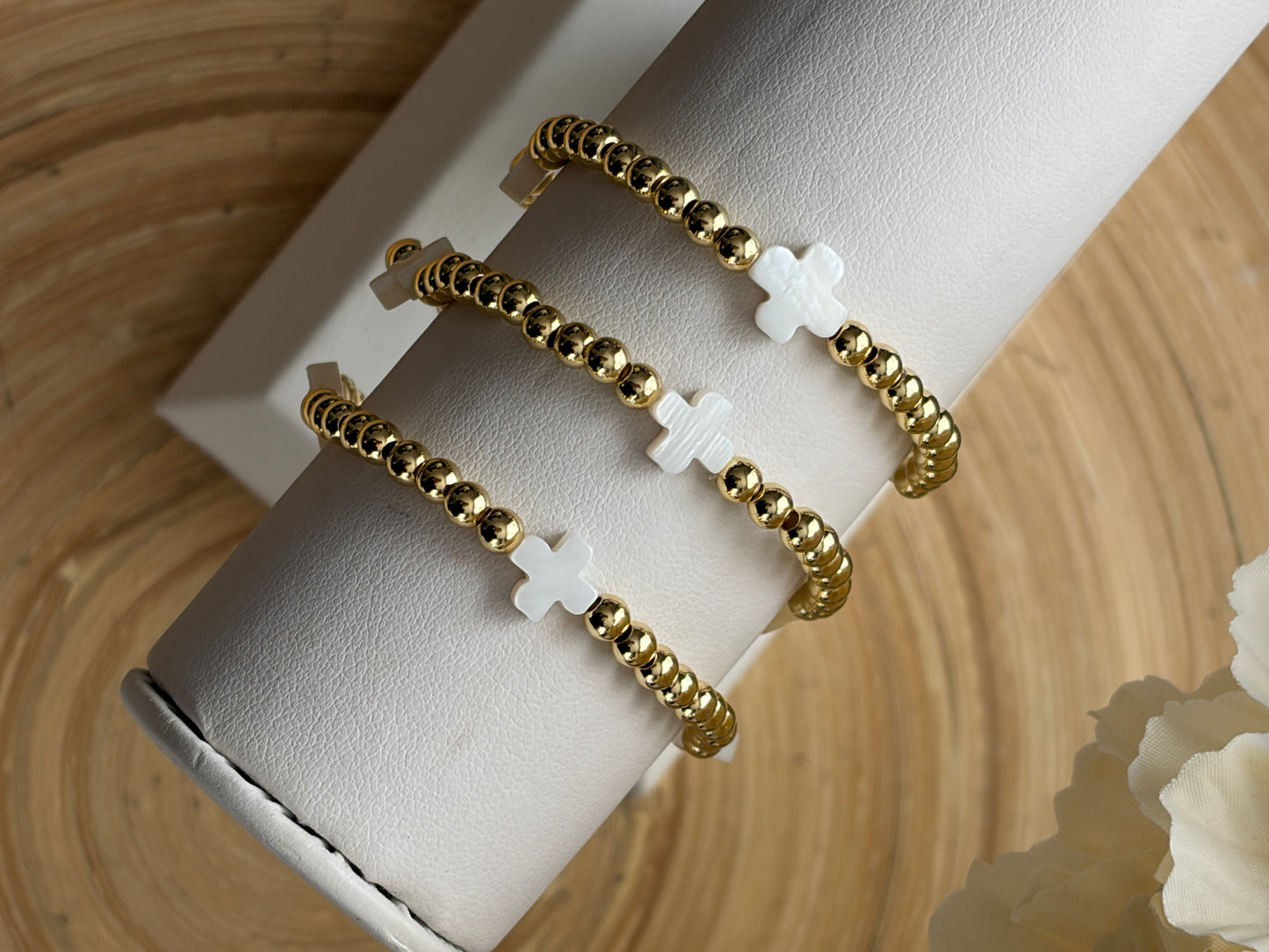 Gold beaded bracelet with white cross charms on a white cylindrical stand against a beige background.