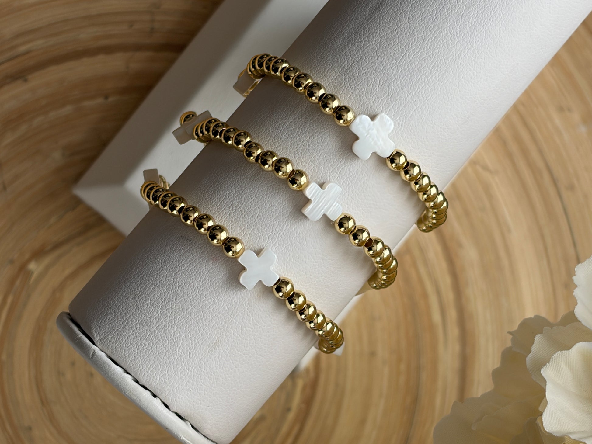 Gold beaded bracelet with white cross charms on a white cylindrical stand against a beige background.