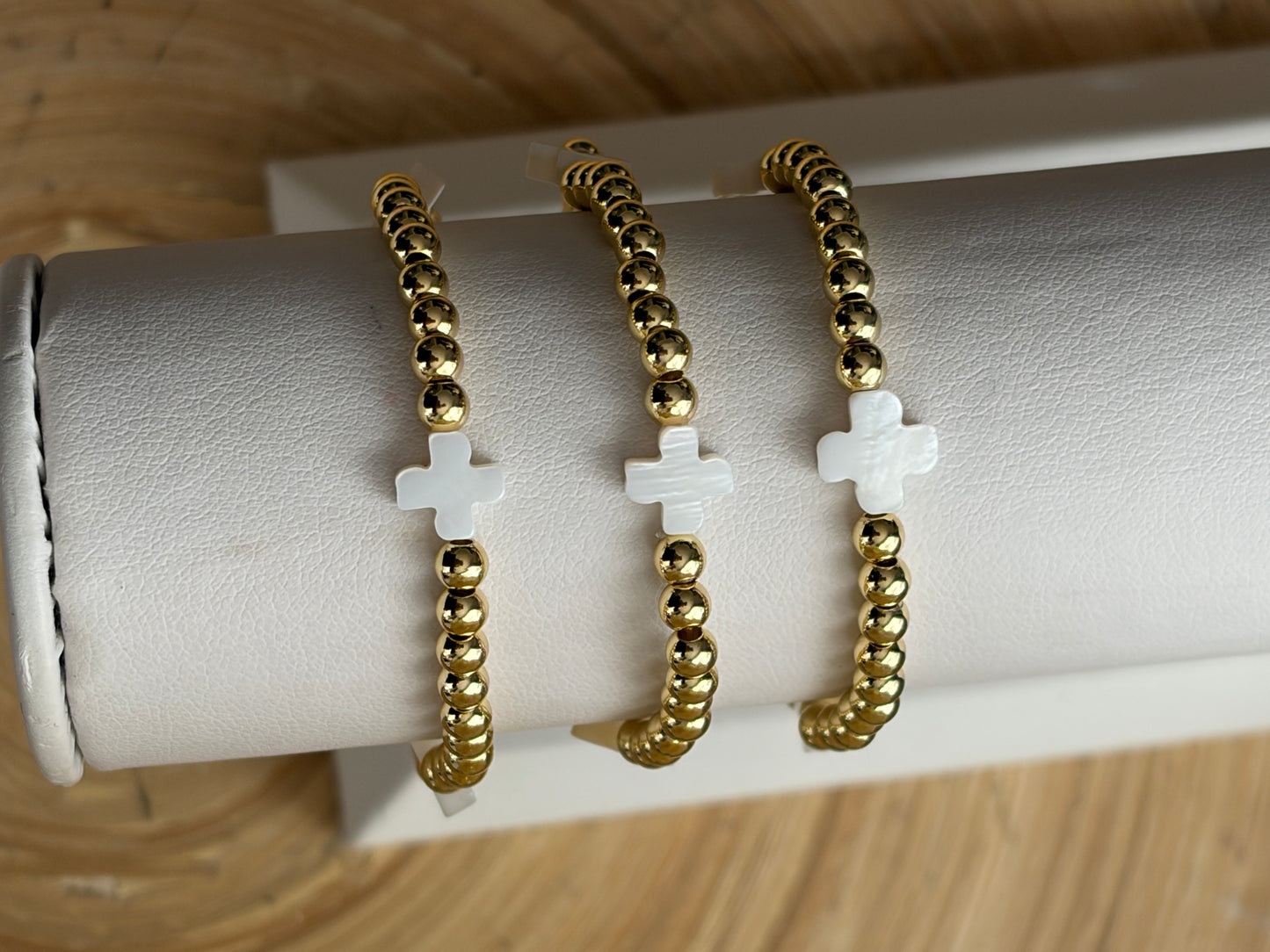 Three gold beaded bracelets with white crosses on a white cushioned stand.