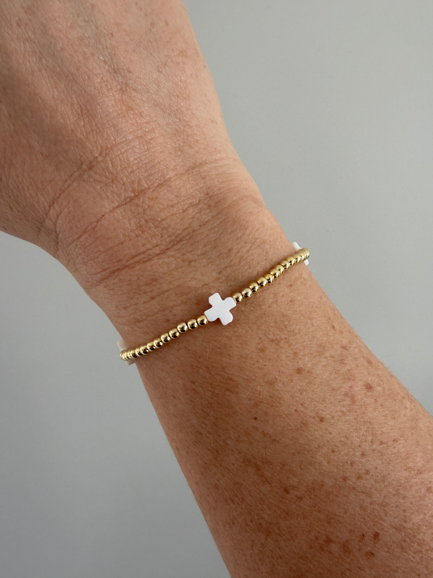 Gold bracelet with a white cross charm on a wrist against a gray background