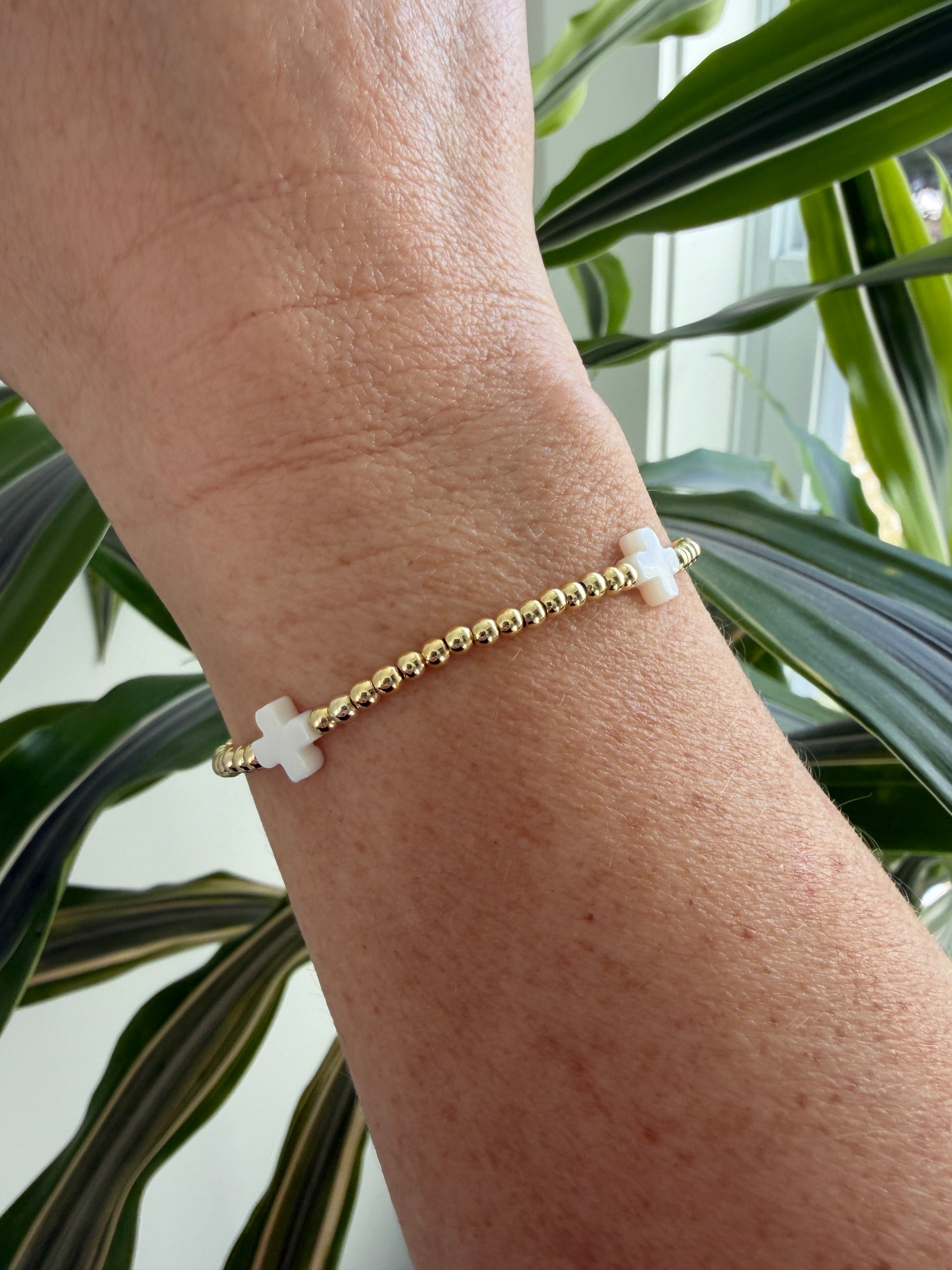 Gold bracelet with a white charm on a wrist, with green plants in the background